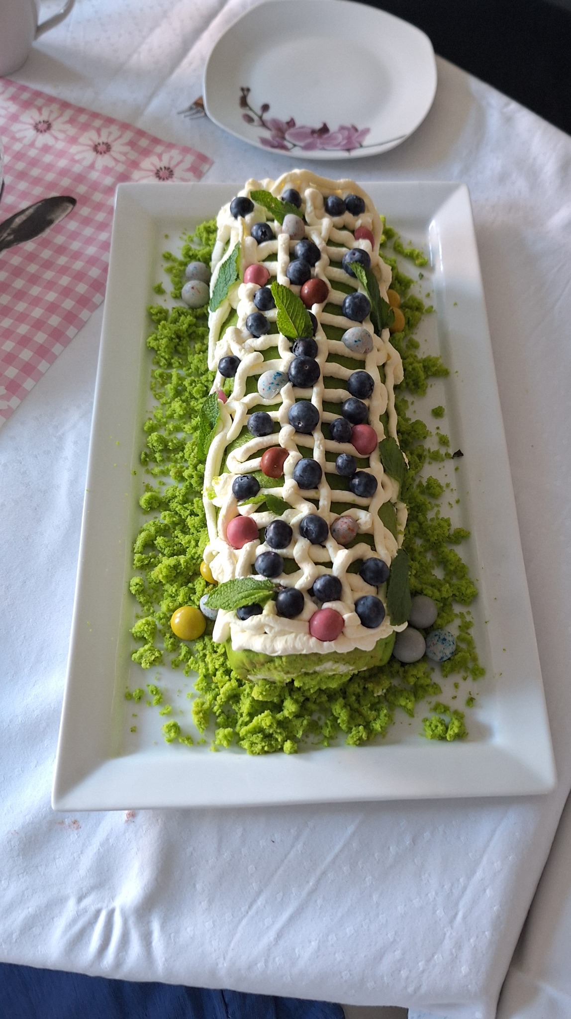 Dekoracyjny torcik na prostokątnym talerzu, ozdobiony kremem, borówkami, miętą i posypką w odcieniach zieleni, leżący na białym obrusie obok talerza z kwiatowym motywem.