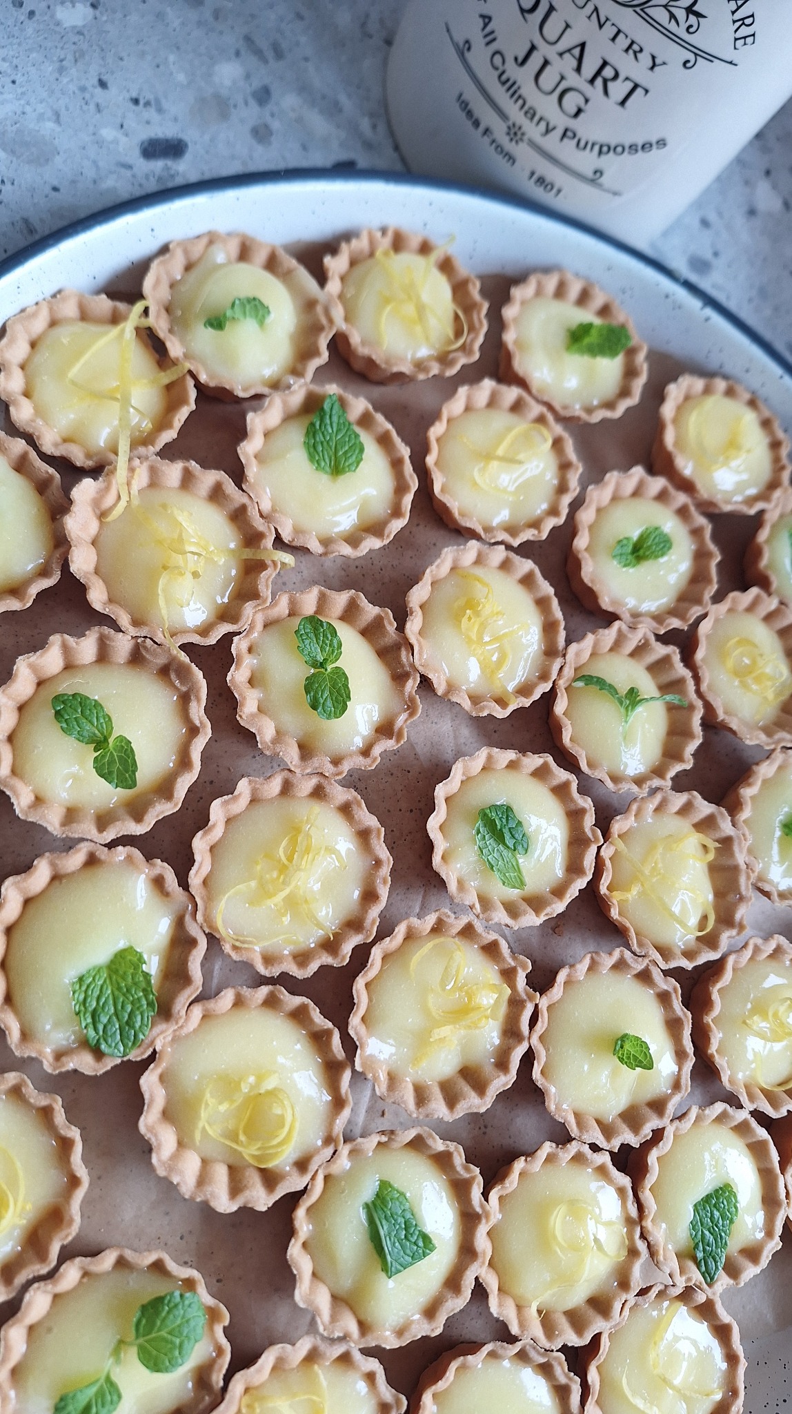 Talerz z mini tartaletkami z kremem cytrynowym, udekorowanymi skórką cytryny i miętą. Obok dzbanek z napisem „Country Quart Jug”.