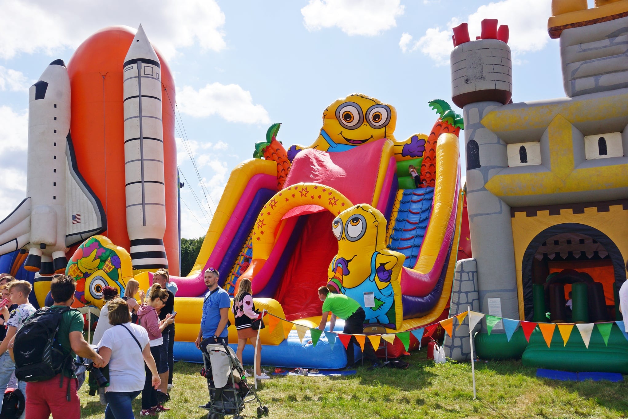 Kolorowy park rozrywki z dmuchanymi atrakcjami: zjeżdżalnie z motywem Minionków, rakieta kosmiczna i zamek. W tle grupa ludzi i błękitne niebo.