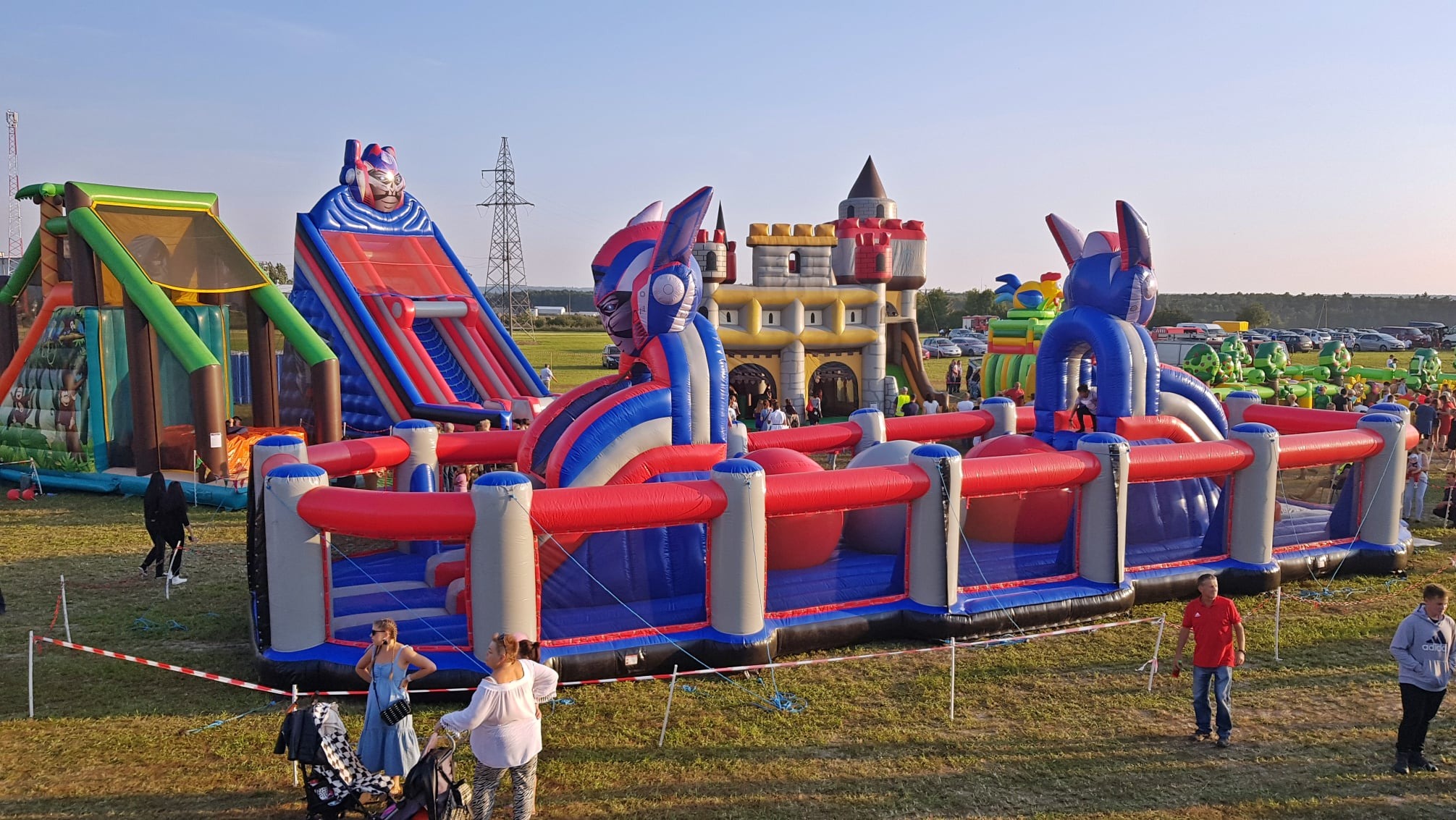 Kolorowe dmuchańce na trawiastym terenie w Janowicach. Widoczny zamek, zjeżdżalnie i tor przeszkód. W tle zaparkowane samochody i ludzie.