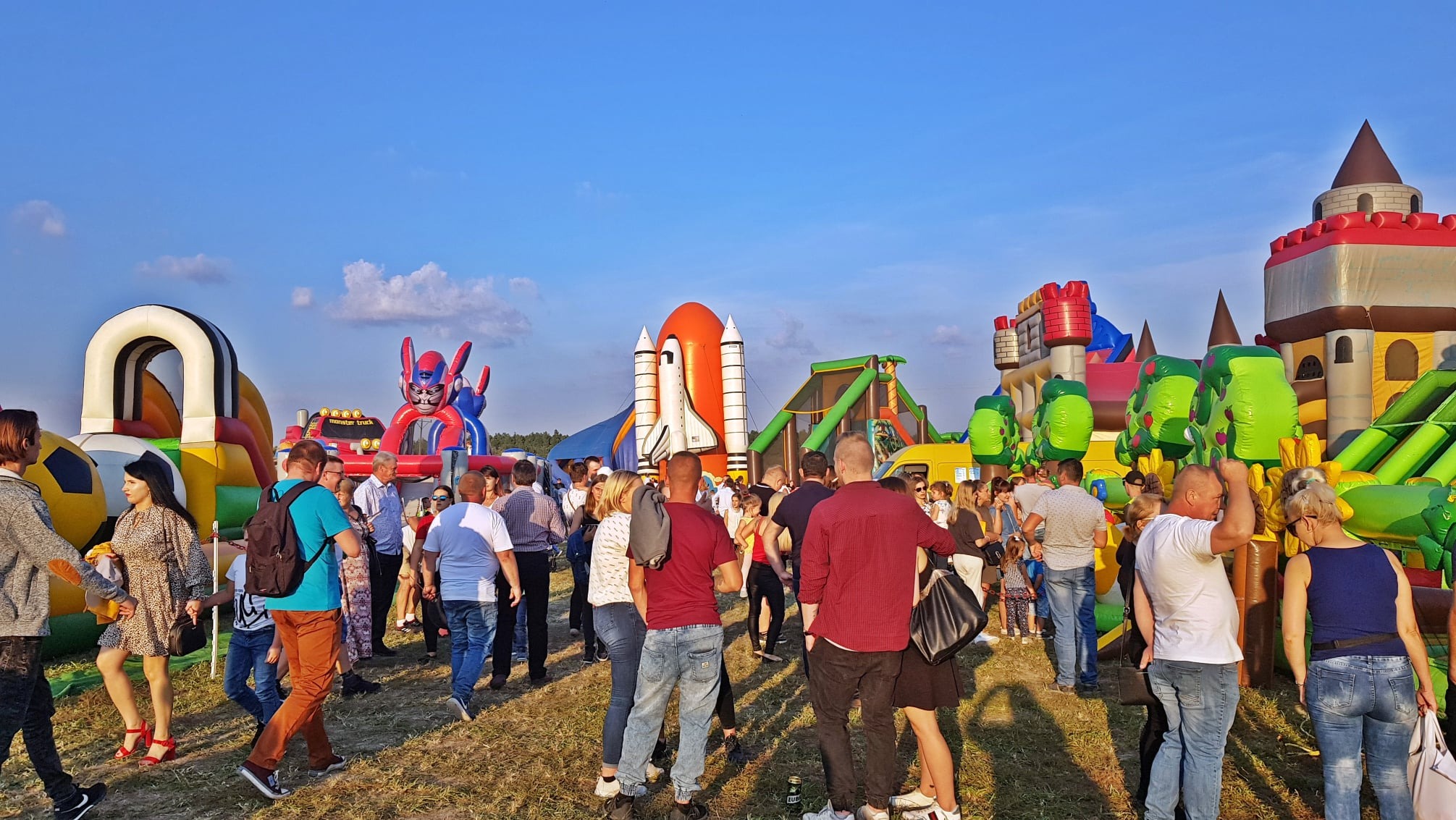 Kolorowe dmuchańce (zamek, rakieta, robot) na otwartym terenie, otoczone grupą ludzi podczas imprezy plenerowej w słoneczny dzień.