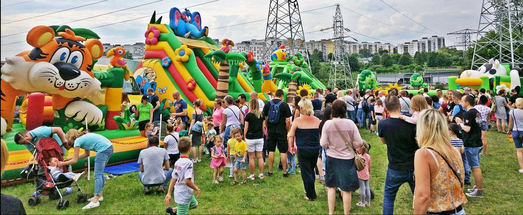 Tłum ludzi na trawie przed dmuchanymi zamkami w kształcie zwierząt, w tle budynki mieszkalne i słupy wysokiego napięcia, impreza plenerowa.
