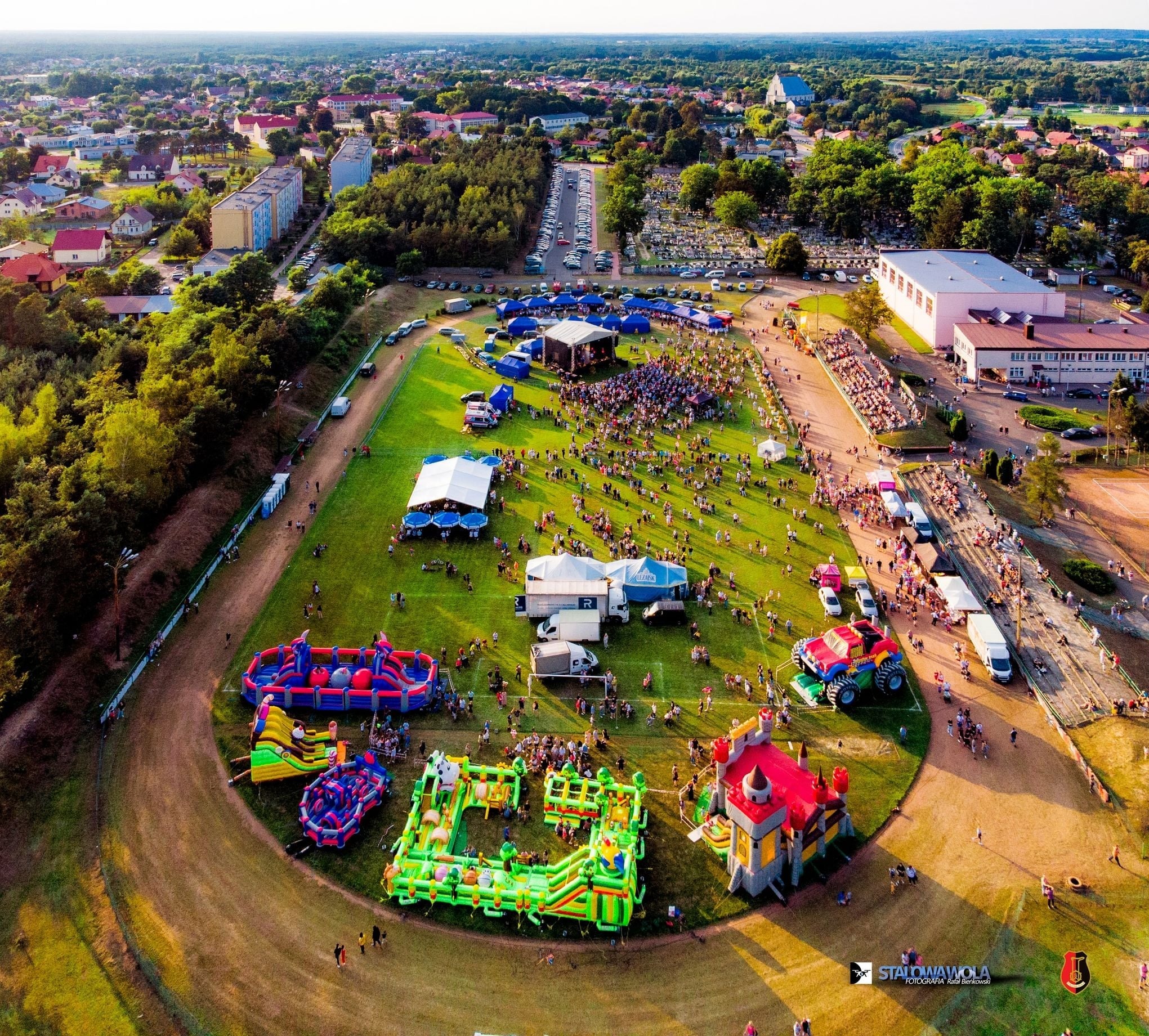 Widok z lotu ptaka na plenerową imprezę z dmuchańcami, sceną i tłumem ludzi. W tle zabudowa miejska i zieleń. Słoneczny dzień.