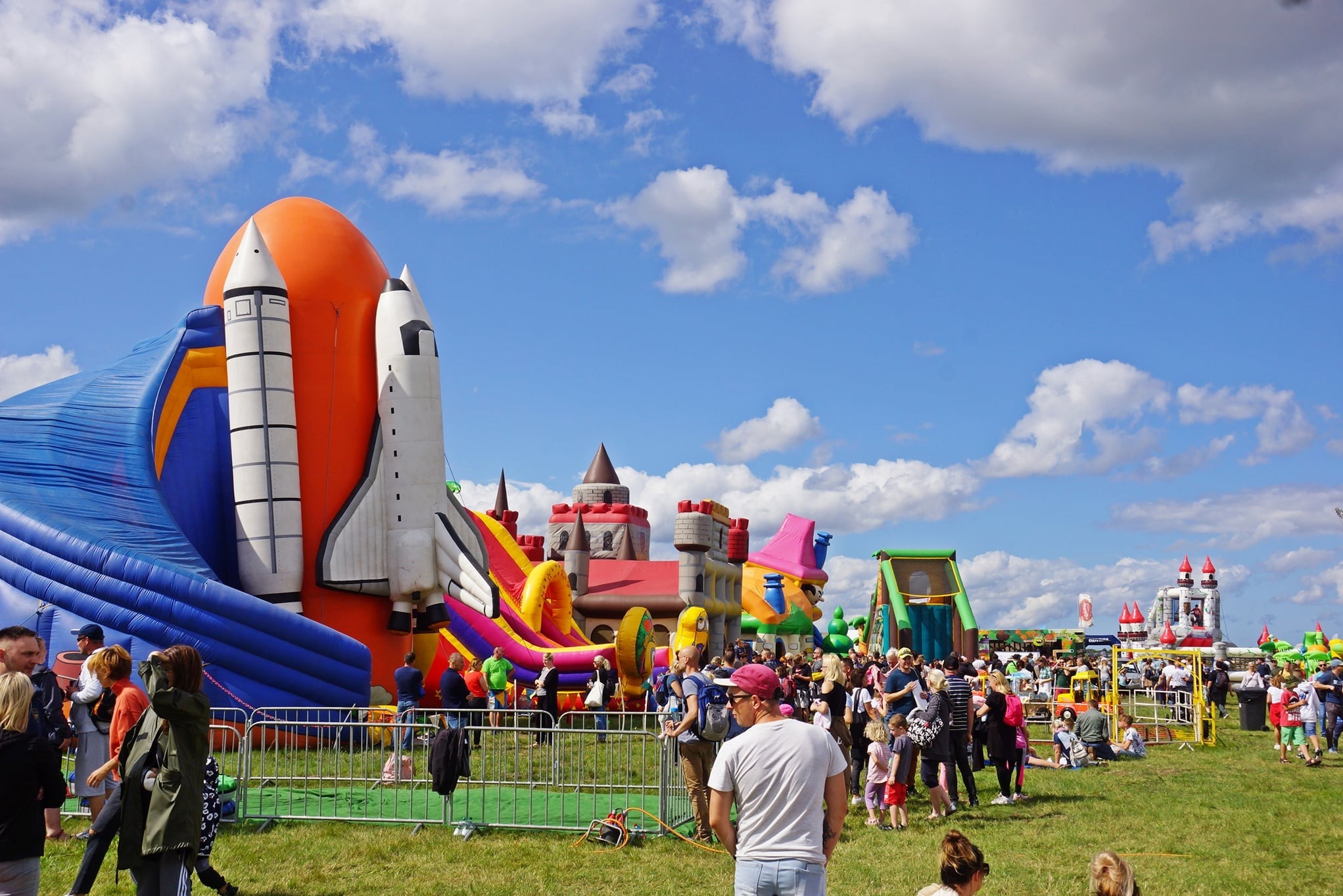 Kolorowe dmuchańce w kształcie rakiet, zamków i zjeżdżalni na zielonej trawie, otoczone przez ludzi w słoneczny dzień. Idealne na imprezy plenerowe.