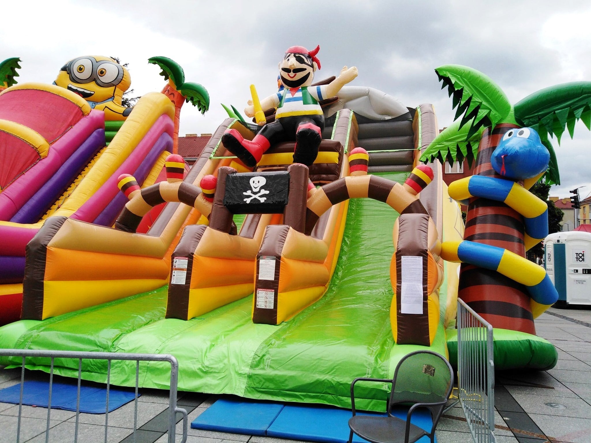 Kolorowy, dmuchany plac zabaw z motywem pirackim, Minionkiem i palmami. Na pierwszym planie krzesło i barierka. W tle widoczne budynki i toaleta przenośna.