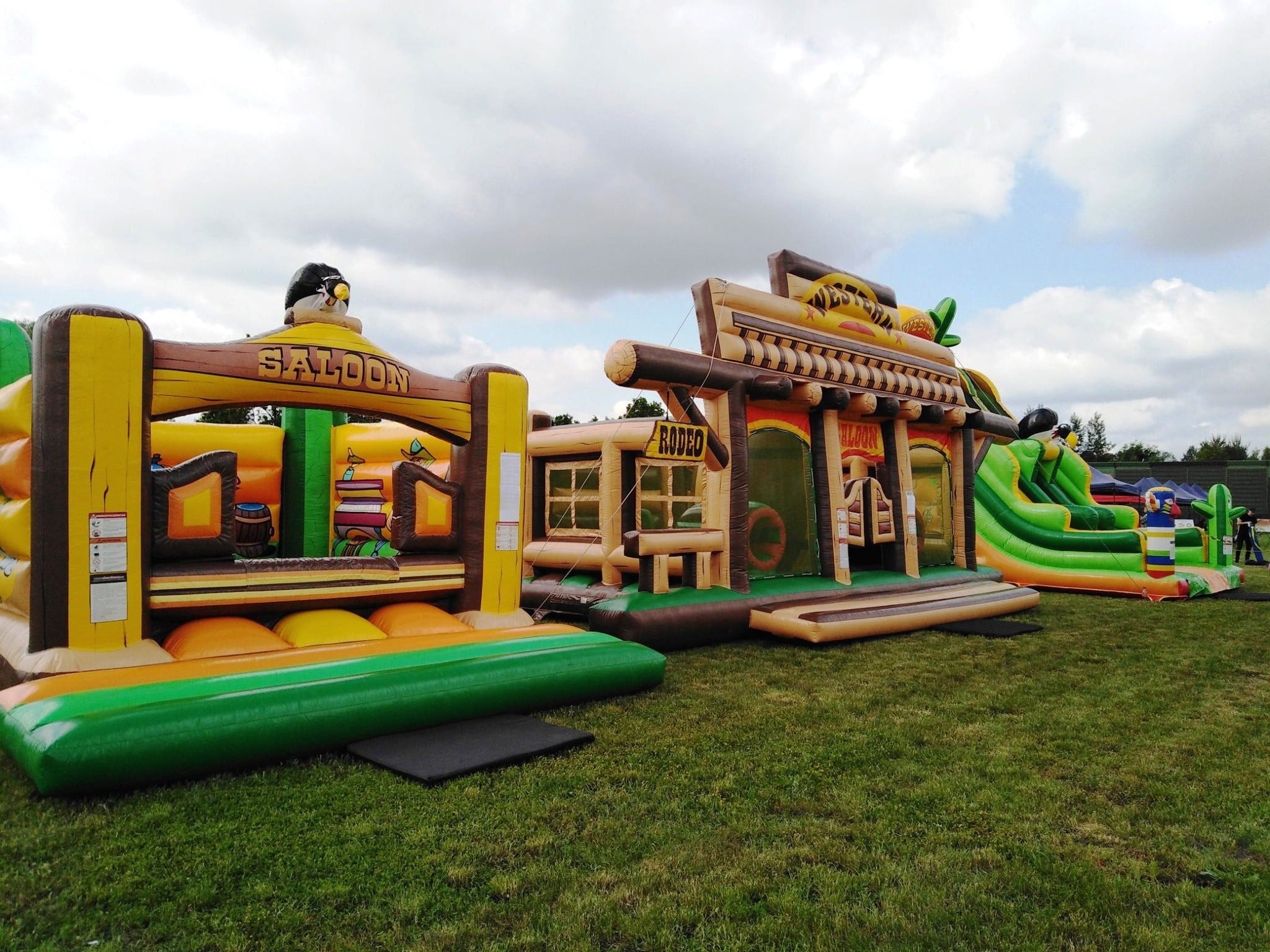 Kompleks dmuchańców w stylu western: Saloon i Rodeo. Kolorowe konstrukcje na trawie, idealne na imprezy plenerowe. Widoczne elementy dekoracyjne.