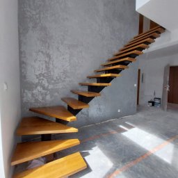 Stolarstwo Projs - Nowoczesne schody drewniane z metalowymi wspornikami na tle ściany o fakturze betonu. Nietypowy kształt stopni i minimalistyczny design.