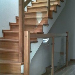Stolarstwo Projs - Nowoczesne schody z jasnego drewna z balustradą ze szkła i drewna, widoczne z dołu. Elementy mocujące balustradę są srebrne. Podłoga wyłożona panelami imitującymi drewno.