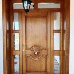 Stolarstwo Projs - Eleganckie drzwi wejściowe z drewna w ciepłym odcieniu, zdobione rzeźbieniami i przeszkleniami, oświetlone wiszącą latarnią, tworzą reprezentacyjne wejście do domu.