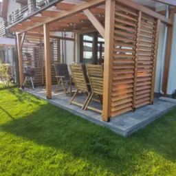 Drewniana pergola z lamelami i oświetleniem na tarasie, z widokiem na dom z balkonem i zadbany trawnik. Dwa krzesła ogrodowe zapraszają do relaksu.