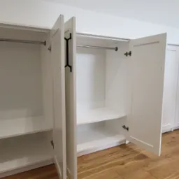 Biała szafa na wymiar z otwartymi drzwiami, widoczne półki i drążek. Na podłodze panele w odcieniach brązu. W tle kolejna szafa z czarnymi uchwytami.