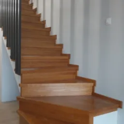 Drewniane schody z ciemnymi metalowymi barierkami, widok z dołu. Stopnie z wyraźnym usłojeniem, pomalowane na jasny brąz. Ściany szare, minimalistyczny design.