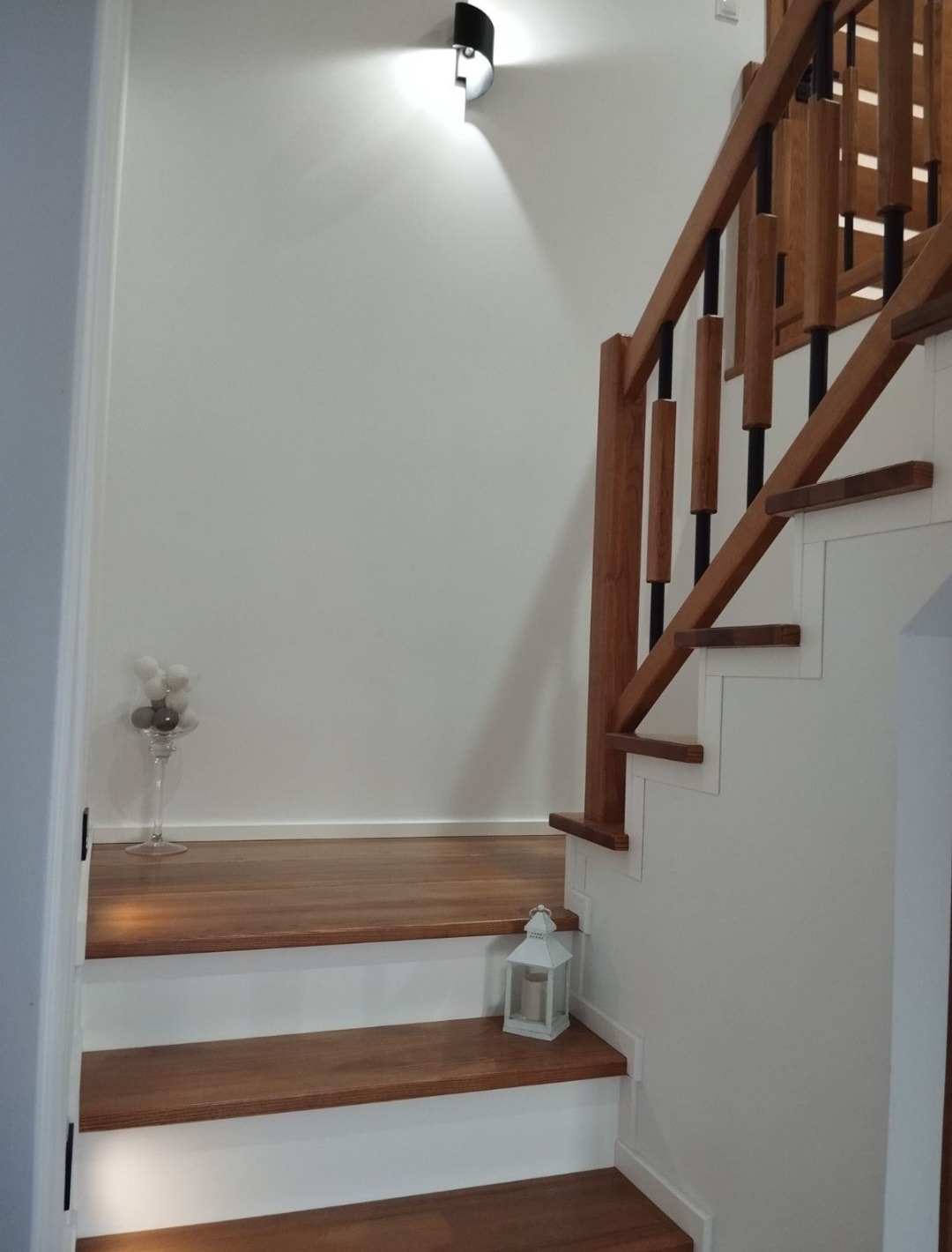 Nowoczesne schody drewniane z białymi podstopnicami, oświetleniem punktowym i dekoracyjną latarenką. Balustrada z drewnianymi i czarnymi elementami.