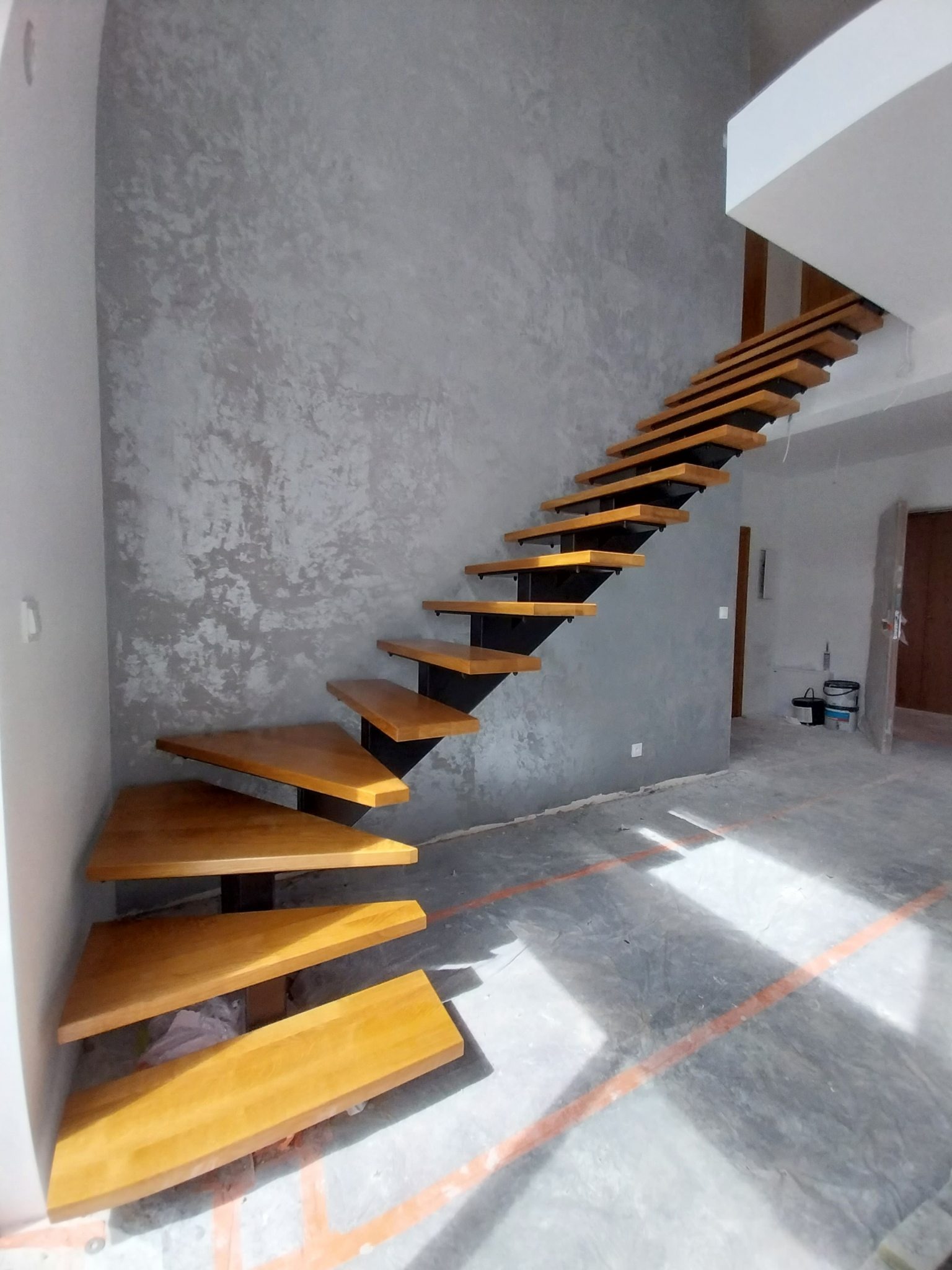 Nowoczesne schody drewniane z metalowymi wspornikami na tle ściany o fakturze betonu. Nietypowy kształt stopni i minimalistyczny design.
