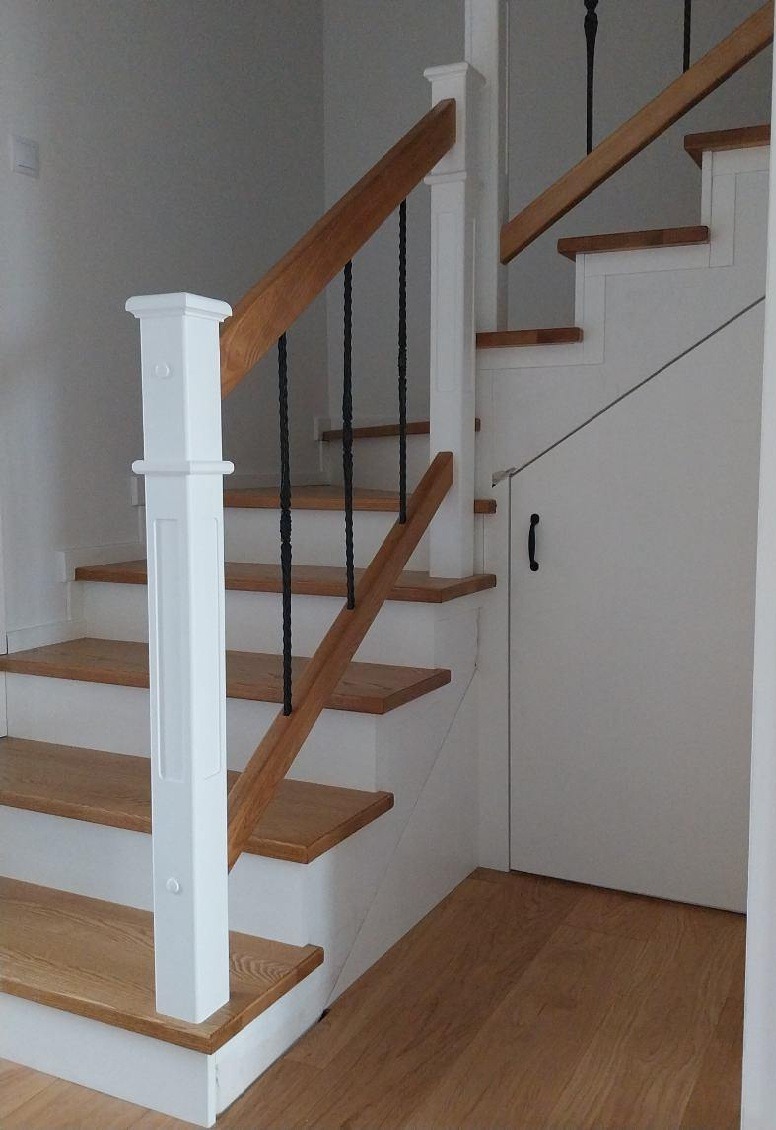 Jasne schody drewniane z białymi elementami i czarnymi, kutymi balustradami. Pod schodami ukryte drzwi. Drewniana podłoga.