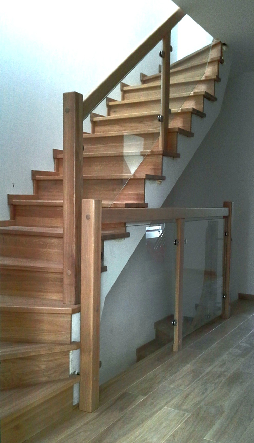 Nowoczesne schody z jasnego drewna z balustradą ze szkła i drewna, widoczne z dołu. Elementy mocujące balustradę są srebrne. Podłoga wyłożona panelami imitującymi drewno.