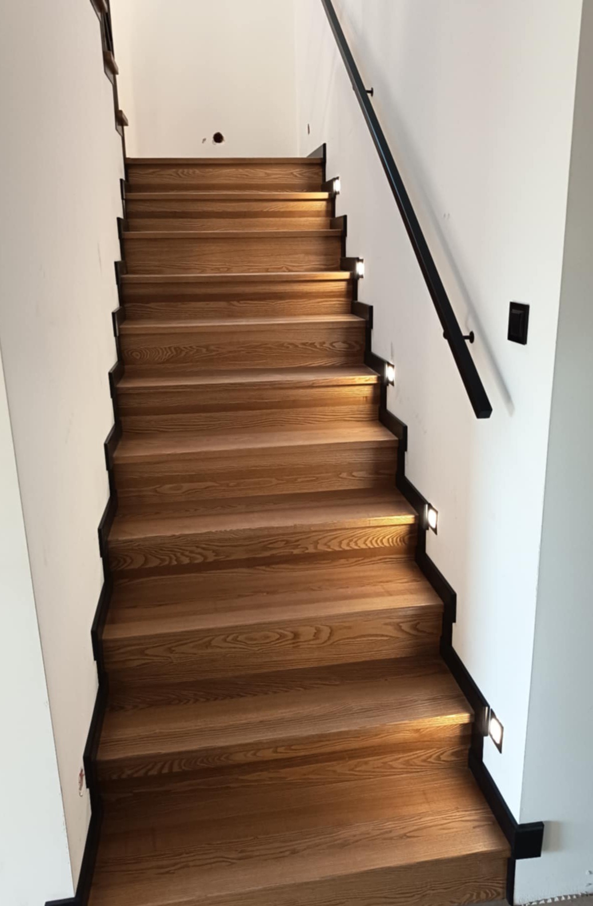 Nowoczesne schody drewniane z podświetleniem LED i czarną balustradą, widok z dołu. Jasne drewno kontrastuje z białymi ścianami, tworząc minimalistyczny design.