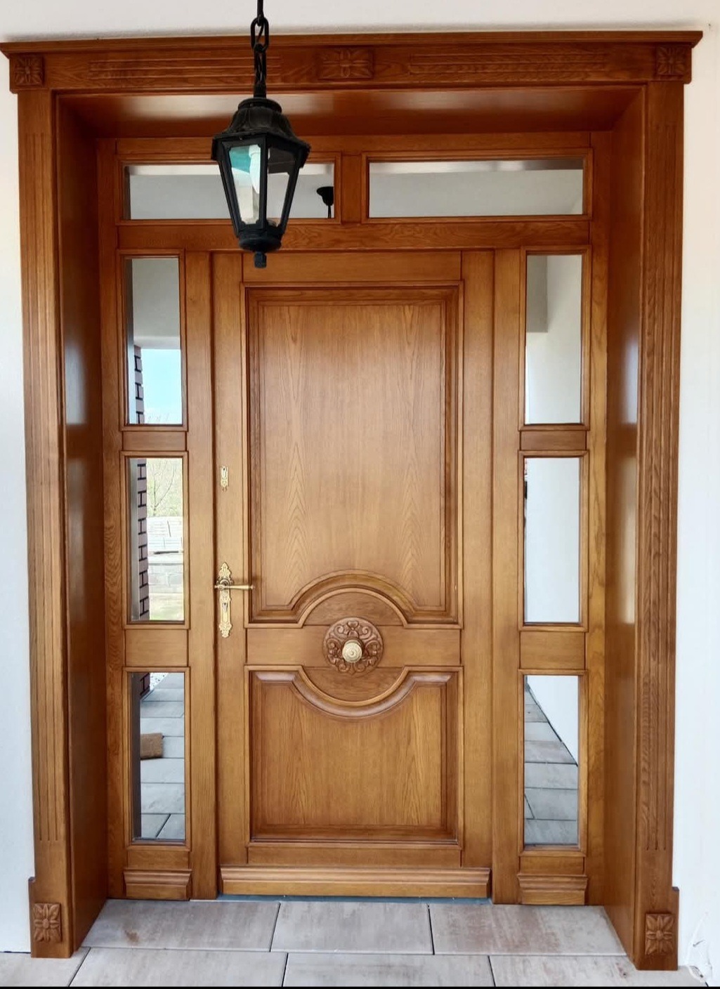 Eleganckie drzwi wejściowe z drewna w ciepłym odcieniu, zdobione rzeźbieniami i przeszkleniami, oświetlone wiszącą latarnią, tworzą reprezentacyjne wejście do domu.