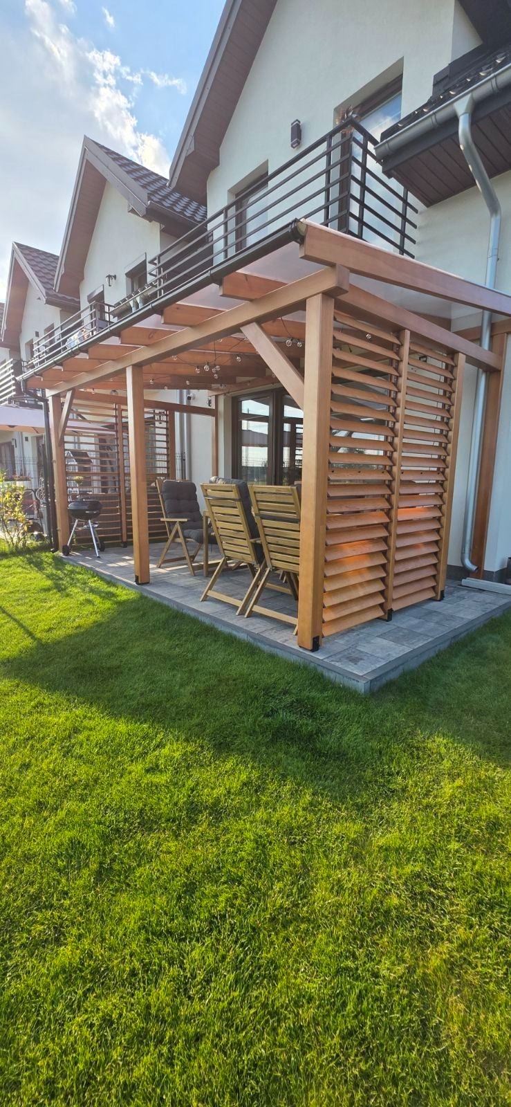 Drewniana pergola z lamelami i oświetleniem na tarasie, z widokiem na dom z balkonem i zadbany trawnik. Dwa krzesła ogrodowe zapraszają do relaksu.