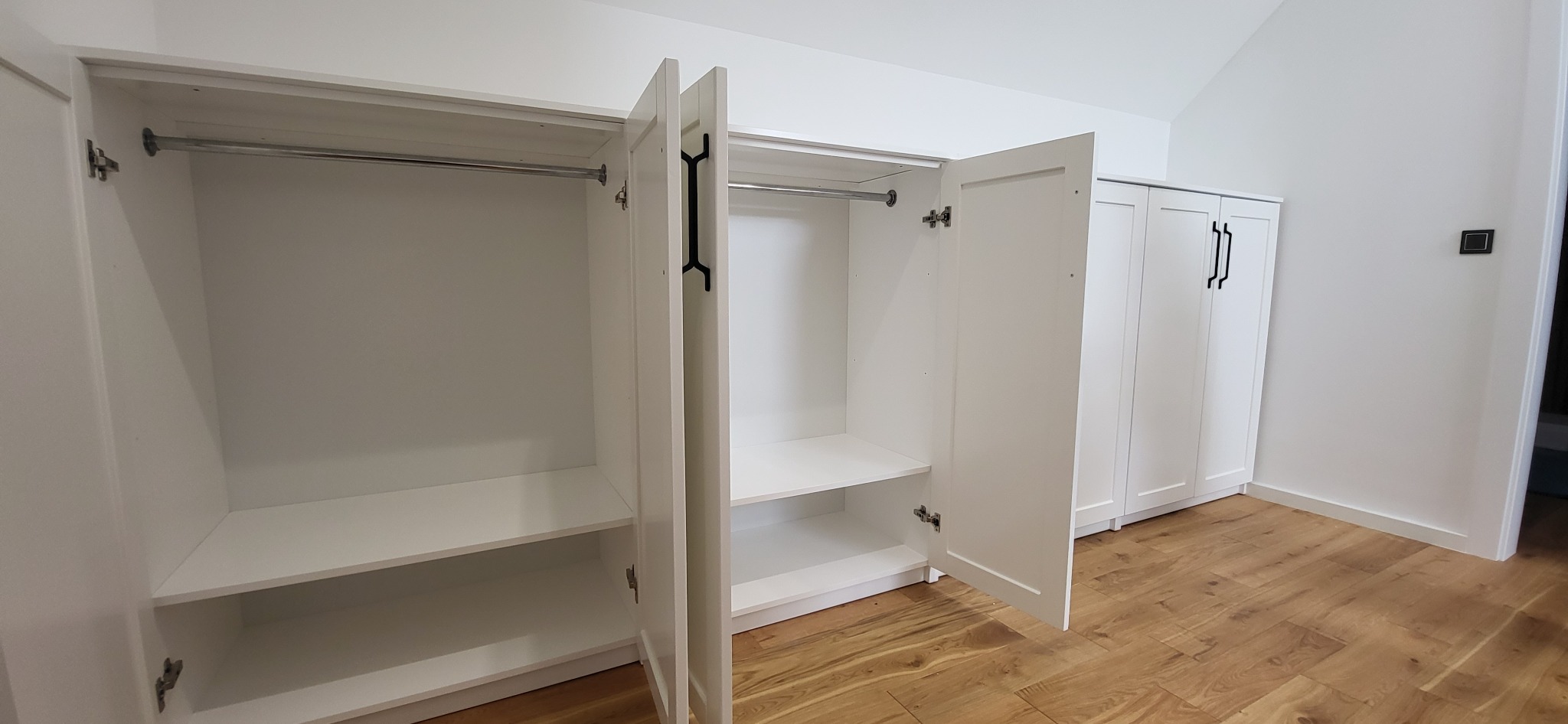 Biała szafa na wymiar z otwartymi drzwiami, widoczne półki i drążek. Na podłodze panele w odcieniach brązu. W tle kolejna szafa z czarnymi uchwytami.