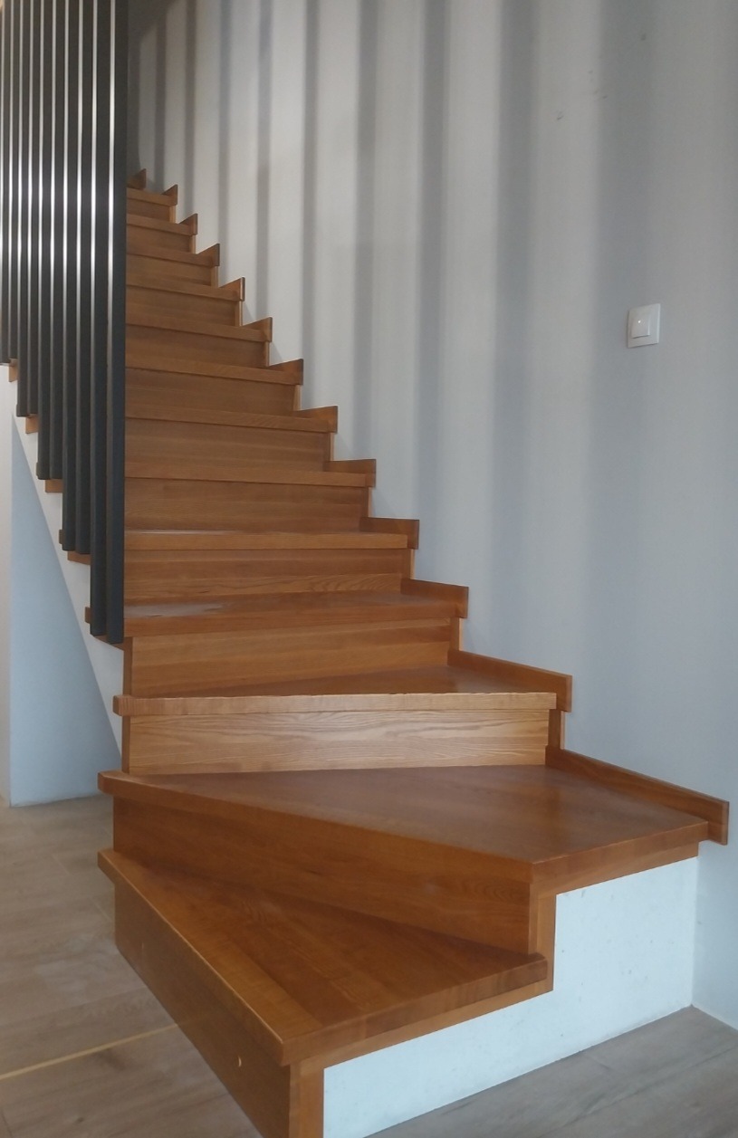 Drewniane schody z ciemnymi metalowymi barierkami, widok z dołu. Stopnie z wyraźnym usłojeniem, pomalowane na jasny brąz. Ściany szare, minimalistyczny design.