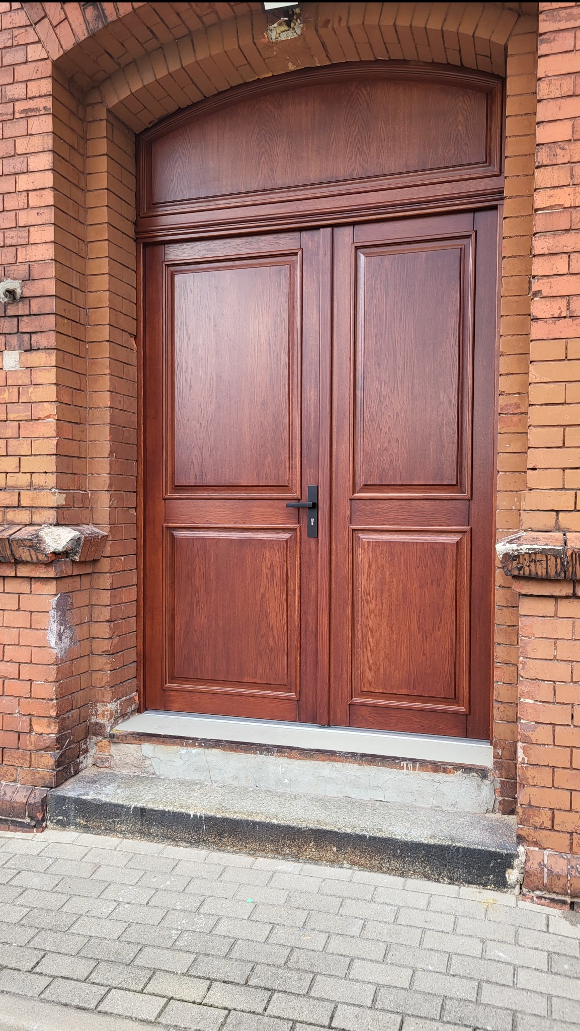Dwuskrzydłowe, ciemnobrązowe drzwi drewniane z czarną klamką, wkomponowane w ceglaną fasadę budynku z łukowym nadprożem. Widoczne detale wykończenia i faktura drewna.