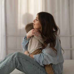 bambolina.photo Natalia Gdak - Kobieta z długimi włosami w dżinsach i koszuli w paski obejmuje małego chłopca na jasnym tle. Delikatne światło i naturalna sceneria.