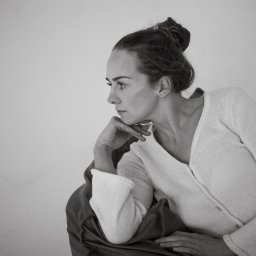 bambolina.photo Natalia Gdak - Monochromatyczne portretowe ujęcie kobiety z upiętymi włosami, opierającej brodę na dłoni, w białym swetrze na tle jasnej ściany. Profil, melancholijny nastrój.