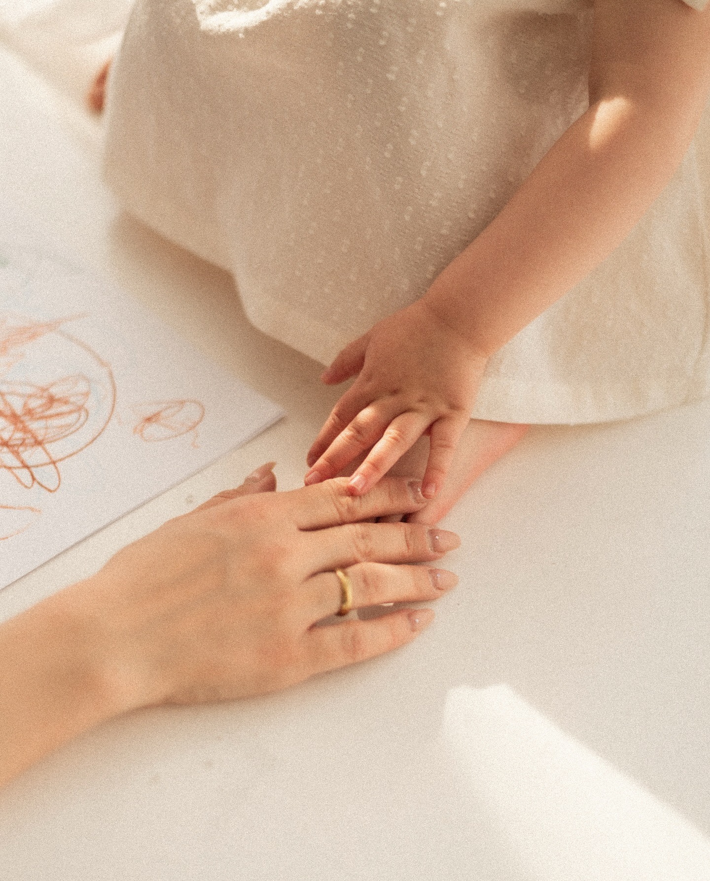 Dłoń matki z złotym pierścionkiem delikatnie dotyka dłoni dziecka na tle rysunku, ciepłe, naturalne światło.