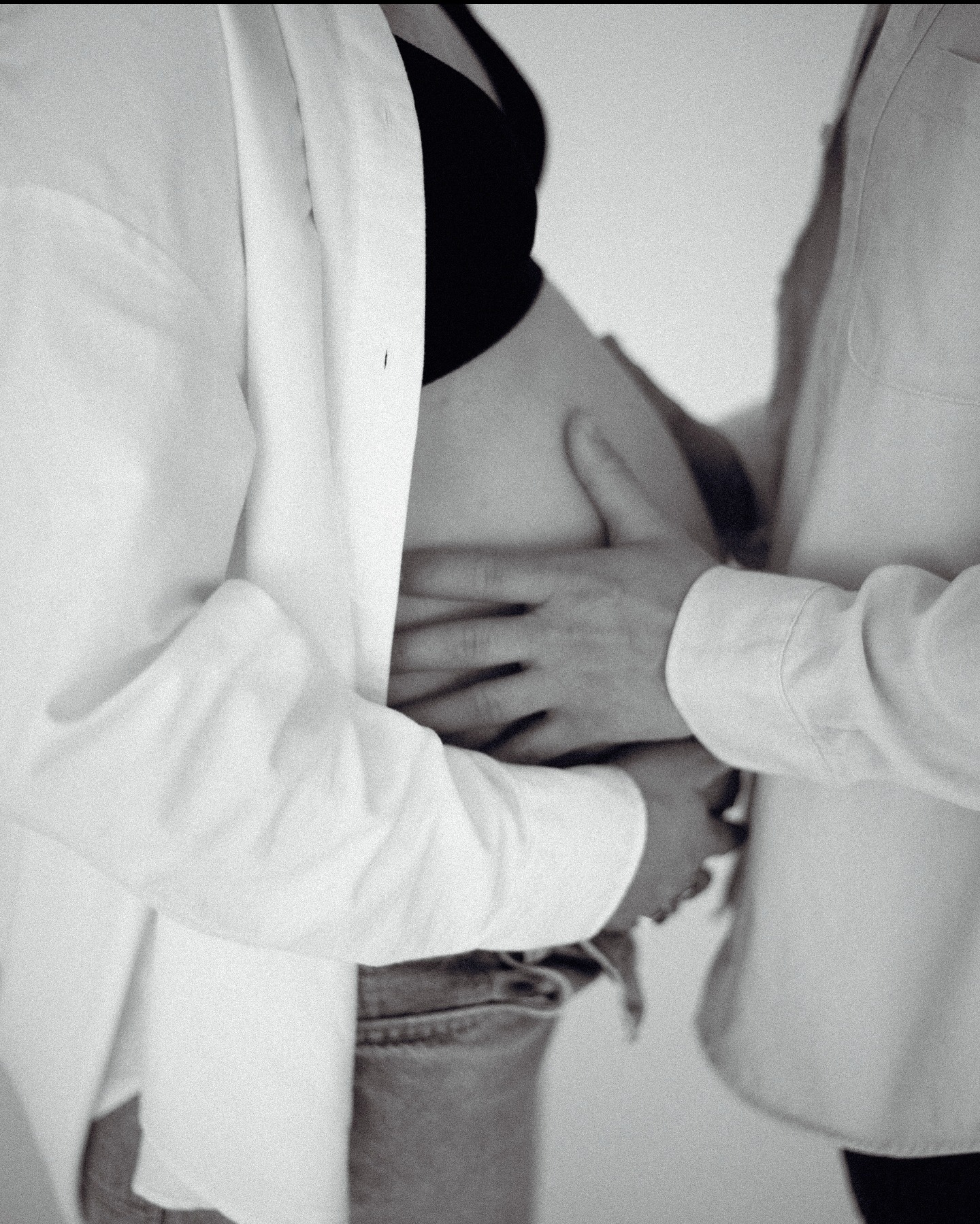 Czarno-białe zbliżenie na ciążowy brzuszek obejmowany przez dwie pary rąk, widoczne białe koszule i dżinsy. Subtelne ujęcie podkreśla intymność i oczekiwanie.
