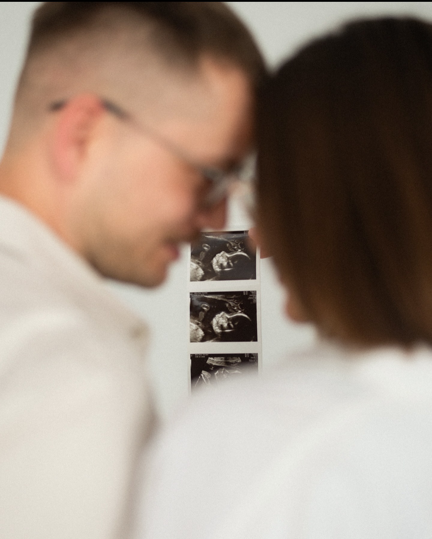 Przyszli rodzice, zamazani na pierwszym planie, patrzą na wydruk USG z boku w jasnym otoczeniu. Mężczyzna w okularach. Delikatne światło.