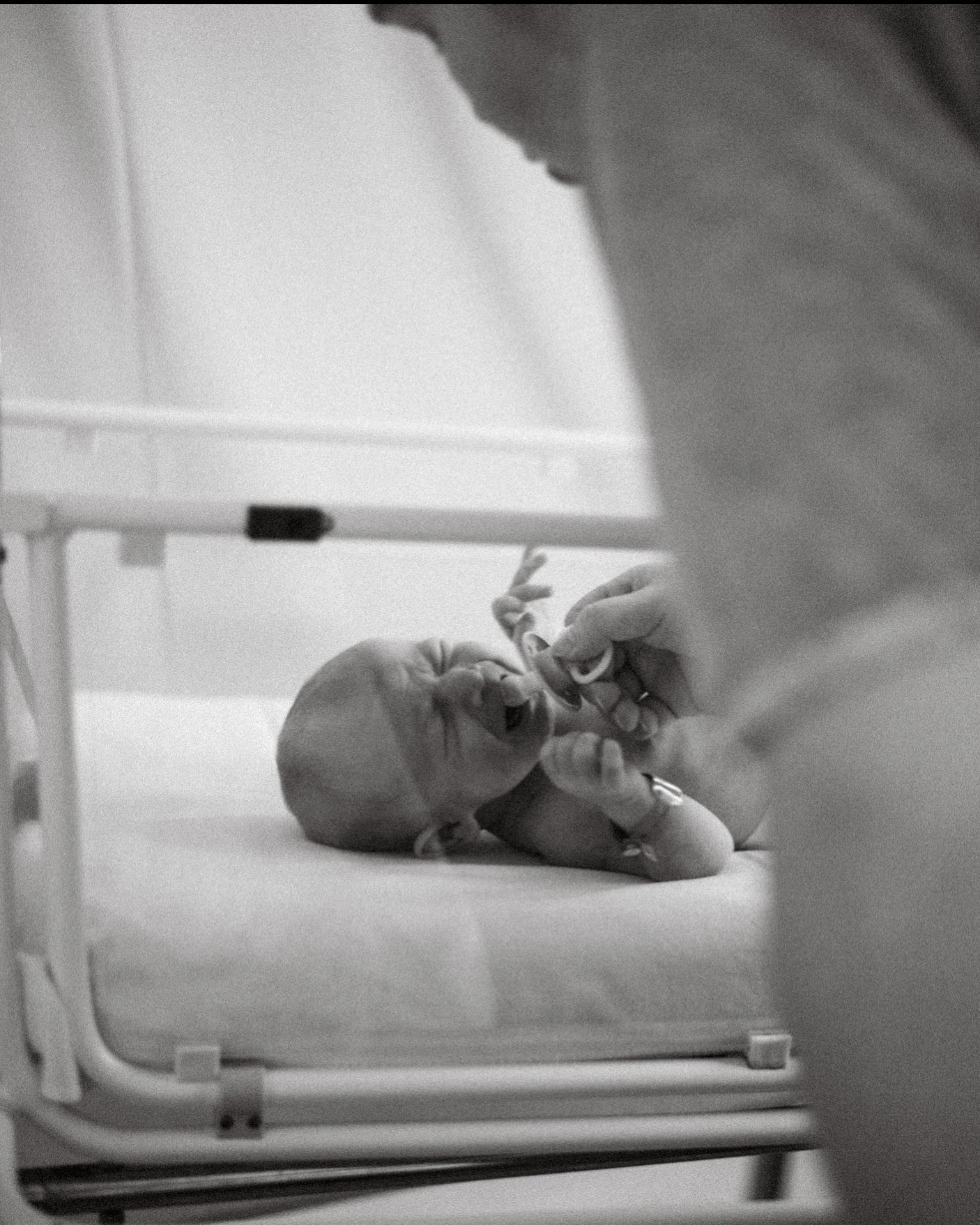 Czarno-białe zdjęcie noworodka leżącego w inkubatorze, z ręką osoby dorosłej podającej smoczek. Dziecko ma zamknięte oczy i otwarte usta. Delikatne, szpitalne oświetlenie.