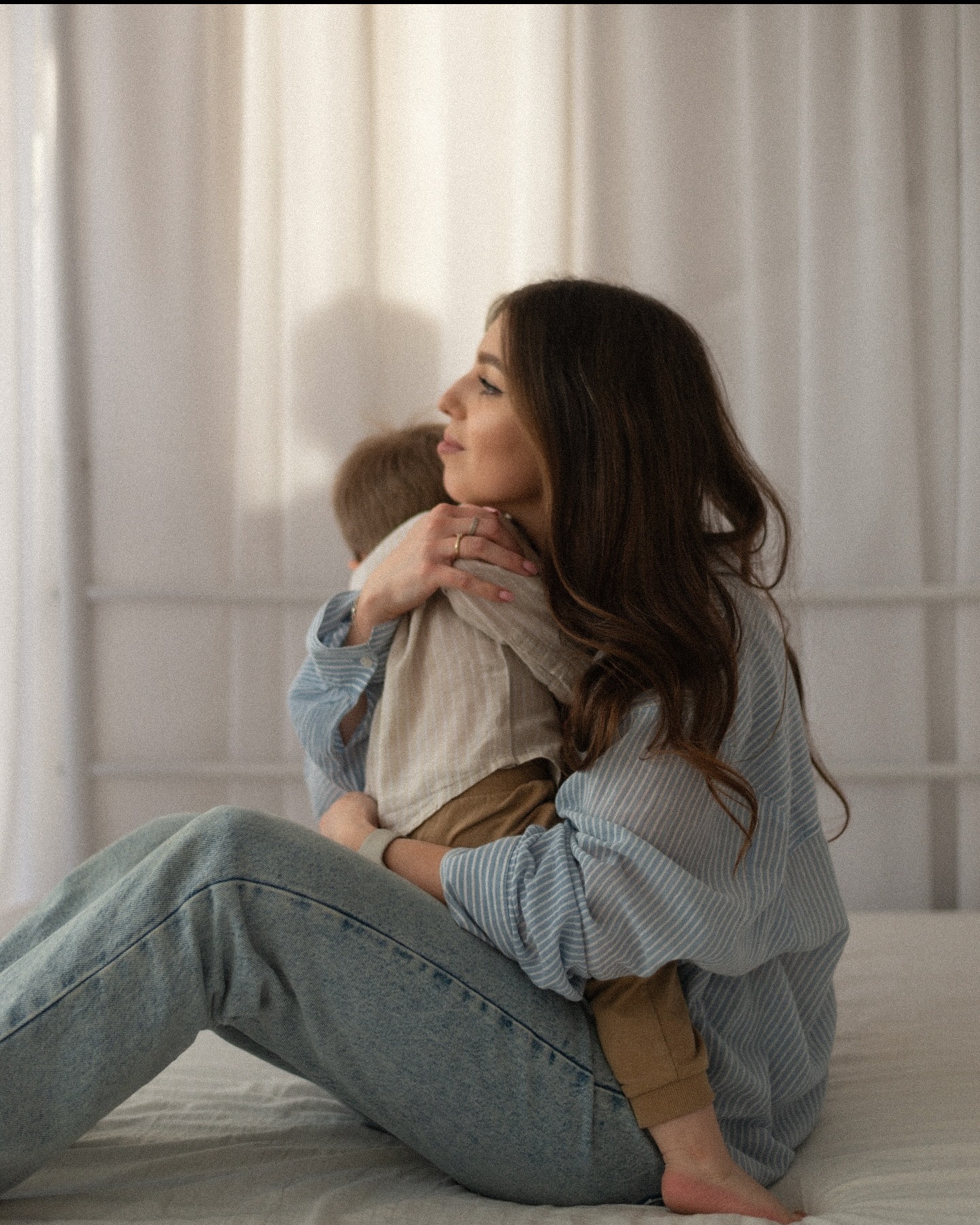 Kobieta z długimi włosami w dżinsach i koszuli w paski obejmuje małego chłopca na jasnym tle. Delikatne światło i naturalna sceneria.