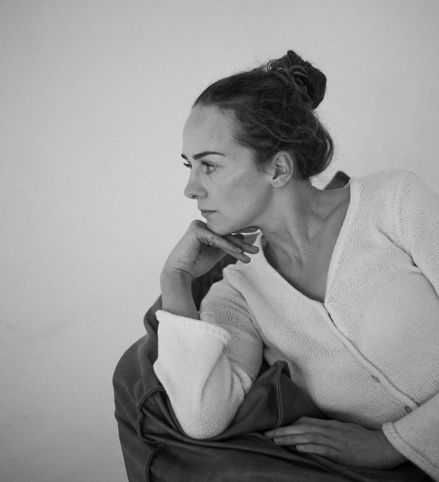 Monochromatyczne portretowe ujęcie kobiety z upiętymi włosami, opierającej brodę na dłoni, w białym swetrze na tle jasnej ściany. Profil, melancholijny nastrój.