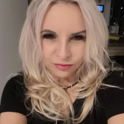 Portret kobiety o blond włosach, z wyrazistym makijażem oczu i delikatnym uśmiechem, ubrana w czarną bluzkę. Ujęcie z bliska, z widocznym tłem wnętrza.