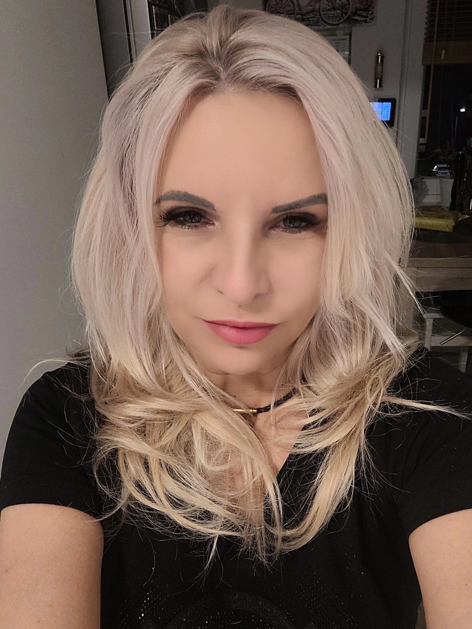 Portret kobiety o blond włosach, z wyrazistym makijażem oczu i delikatnym uśmiechem, ubrana w czarną bluzkę. Ujęcie z bliska, z widocznym tłem wnętrza.