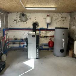 Zainstalowany piec Defro Evopell z zasobnikiem Trinity i instalacją CO w Młyniec. Widoczne rury w kolorach czerwonym i niebieskim. Ściany z widoczną strukturą betonu.