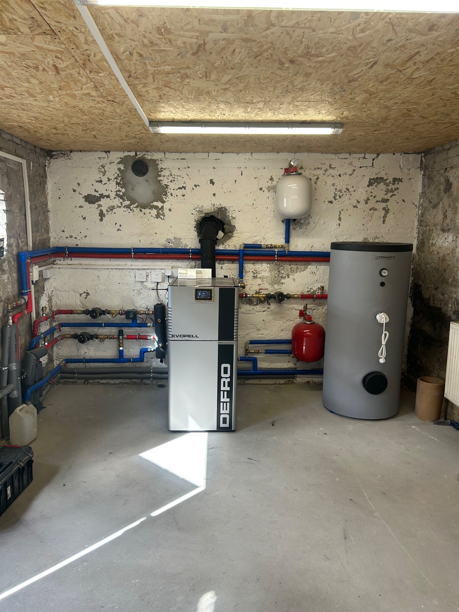 Zainstalowany piec Defro Evopell z zasobnikiem Trinity i instalacją CO w Młyniec. Widoczne rury w kolorach czerwonym i niebieskim. Ściany z widoczną strukturą betonu.