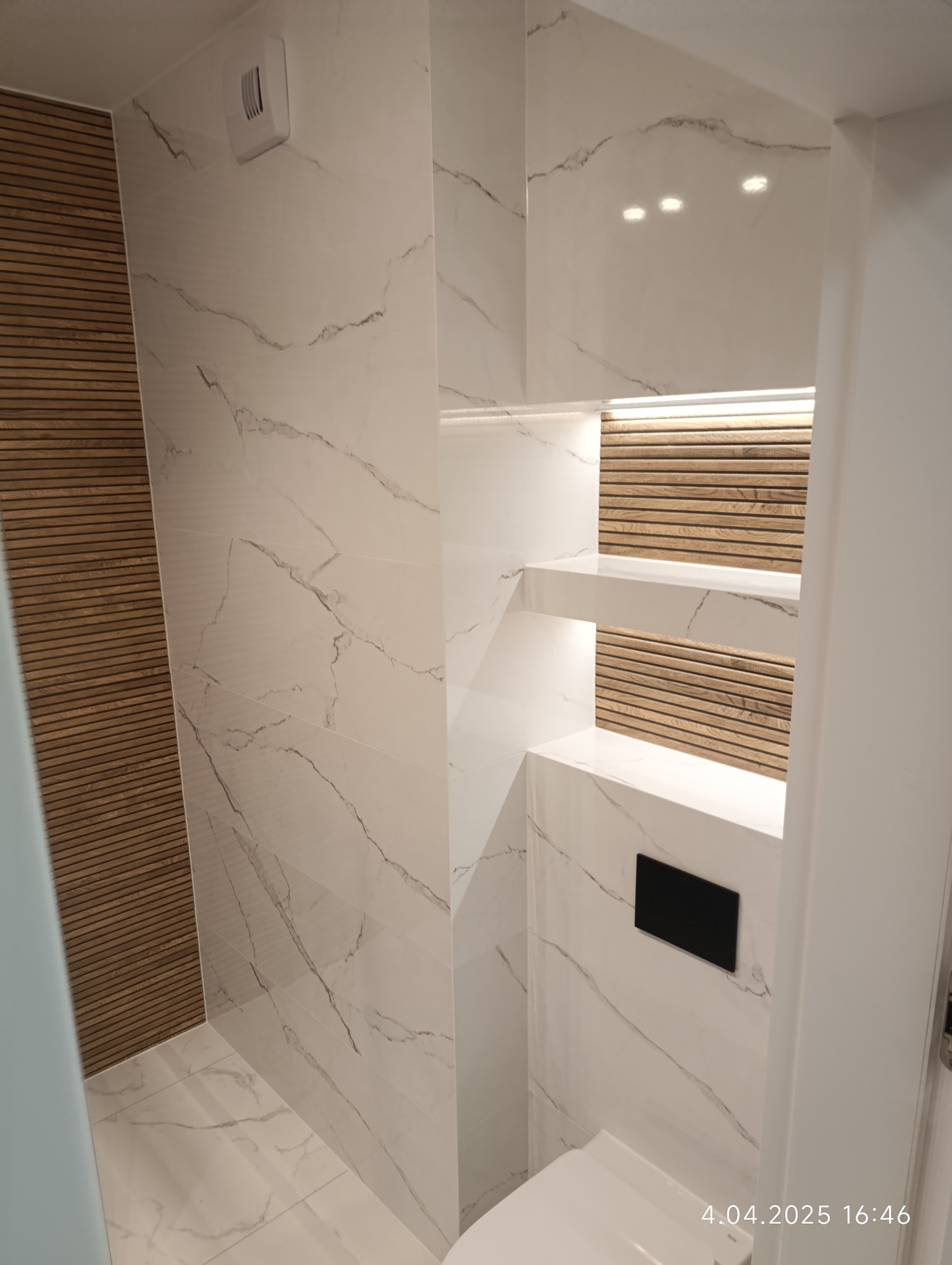Nowoczesna łazienka z białymi płytkami imitującymi marmur, drewnianymi akcentami i podświetlanymi półkami wnękowymi. Widoczny fragment toalety i minimalistyczny design.