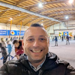 Łukasz Siciarski Zattarra Private chauffeur - Uśmiechnięty mężczyzna na tle lodowiska w hali. W tle osoby jeżdżące na łyżwach i banery reklamowe. Nietypowe ujęcie z bliska, podkreślające atmosferę miejsca.