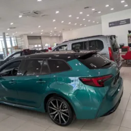 Turkusowa Toyota Corolla kombi w salonie samochodowym, z widocznymi detalami karoserii i felg, na tle jasnego wnętrza salonu z innymi pojazdami.