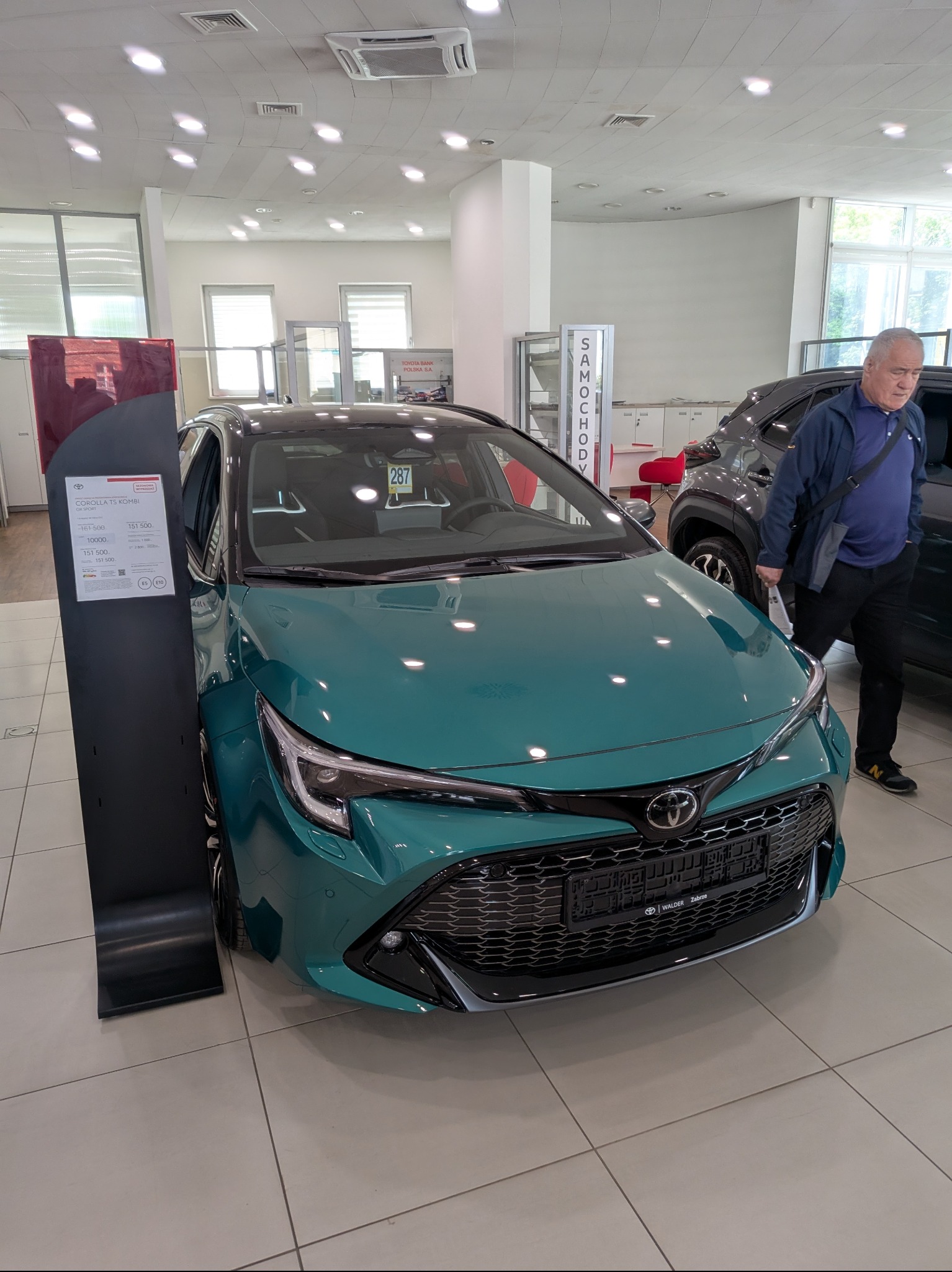 Turkusowa Toyota Corolla TS Kombi GM Sport w salonie samochodowym, widok z przodu. Obok cennik i mężczyzna oglądający inny model. Jasne wnętrze salonu.
