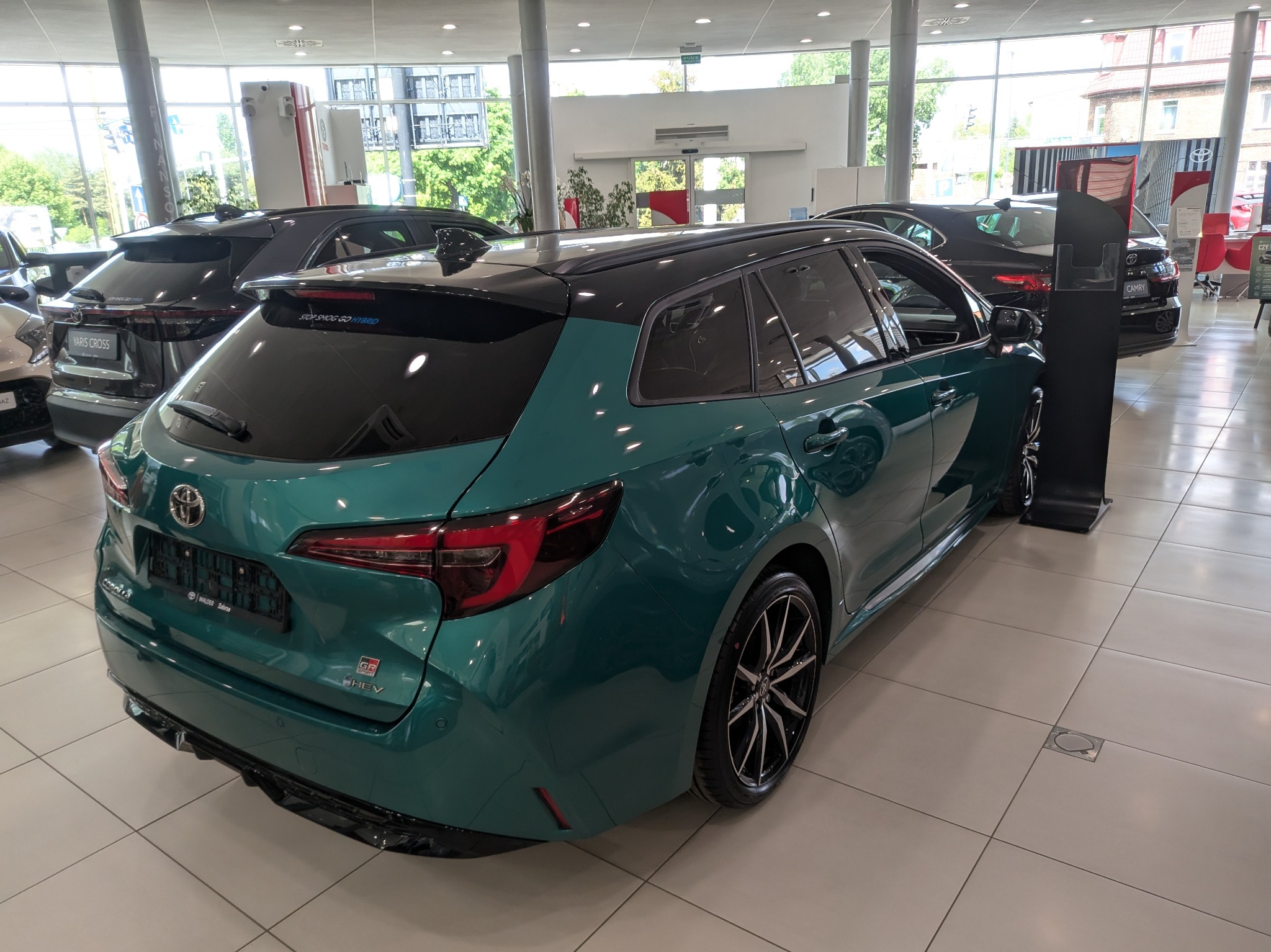Szmaragdowa Toyota Corolla GR HEV kombi w salonie, z czarnym dachem i felgami, widziana z tyłu pod kątem. W tle inne nowe auta i jasne, przestronne wnętrze salonu.