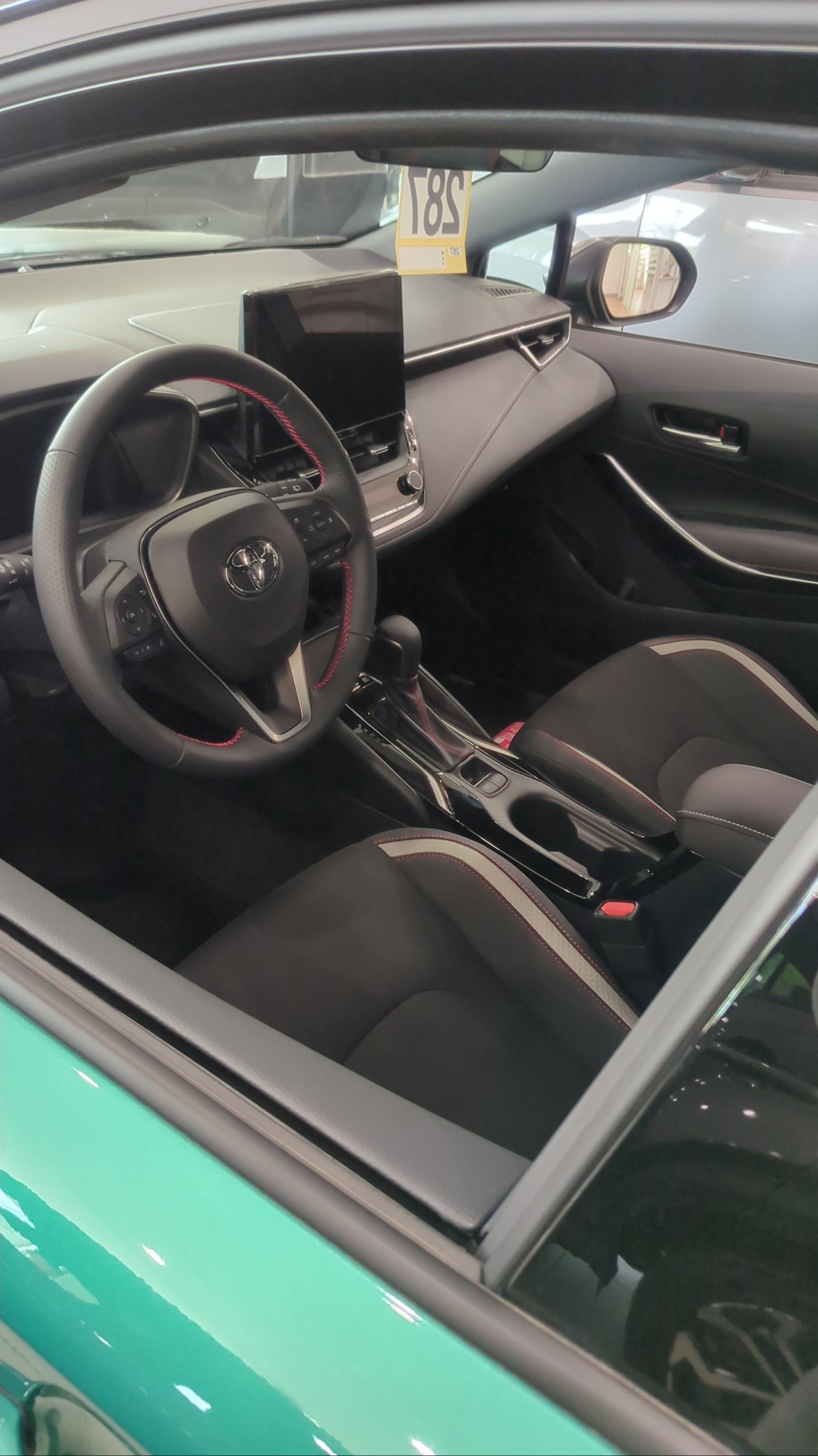 Wnętrze zielonej Toyoty Corolli: kierownica z czerwonymi przeszyciami, czarne fotele z białymi paskami, ekran dotykowy. Widok z zewnątrz przez szybę.