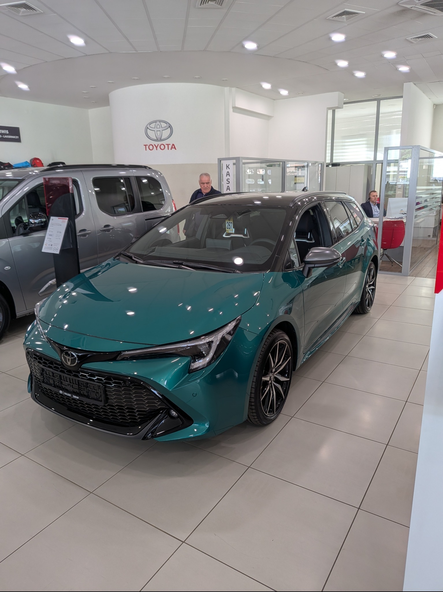 Szmaragdowa Toyota Corolla kombi w salonie, z czarnym dachem i felgami. W tle logo Toyota i inne auta. Dobre oświetlenie, czyste wnętrze salonu.