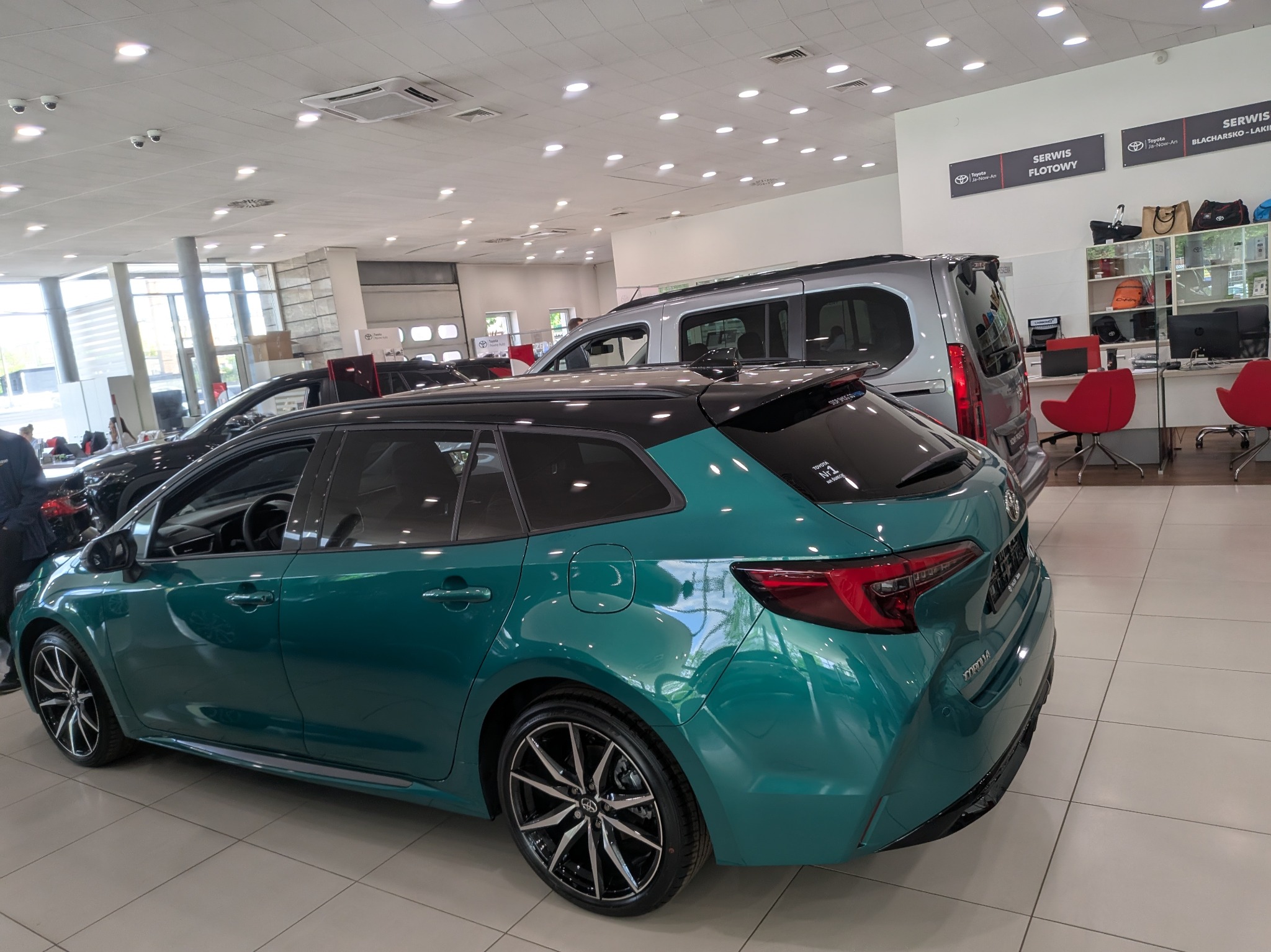 Turkusowa Toyota Corolla kombi w salonie samochodowym, z widocznymi detalami karoserii i felg, na tle jasnego wnętrza salonu z innymi pojazdami.