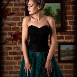 Beata Smołka Fotografia - Elegancka kobieta w czarno-zielonej sukni pozuje na tle ceglanej ściany z obrazami, prezentując subtelny makijaż i biżuterię. Stonowane oświetlenie podkreśla detale.