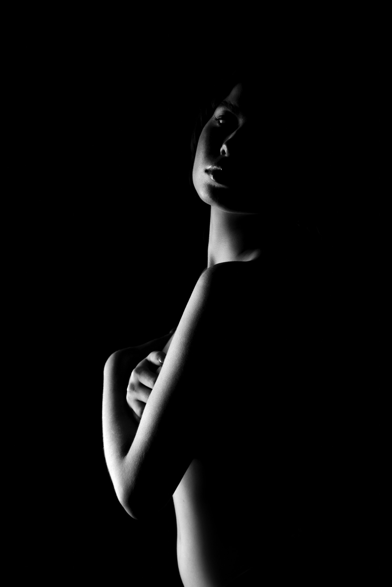 Czarno-biały, artystyczny portret kobiety z ramionami skrzyżowanymi na piersiach, oświetlony w stylu low-key z mocnym kontrastem światła i cienia na gładkiej skórze.