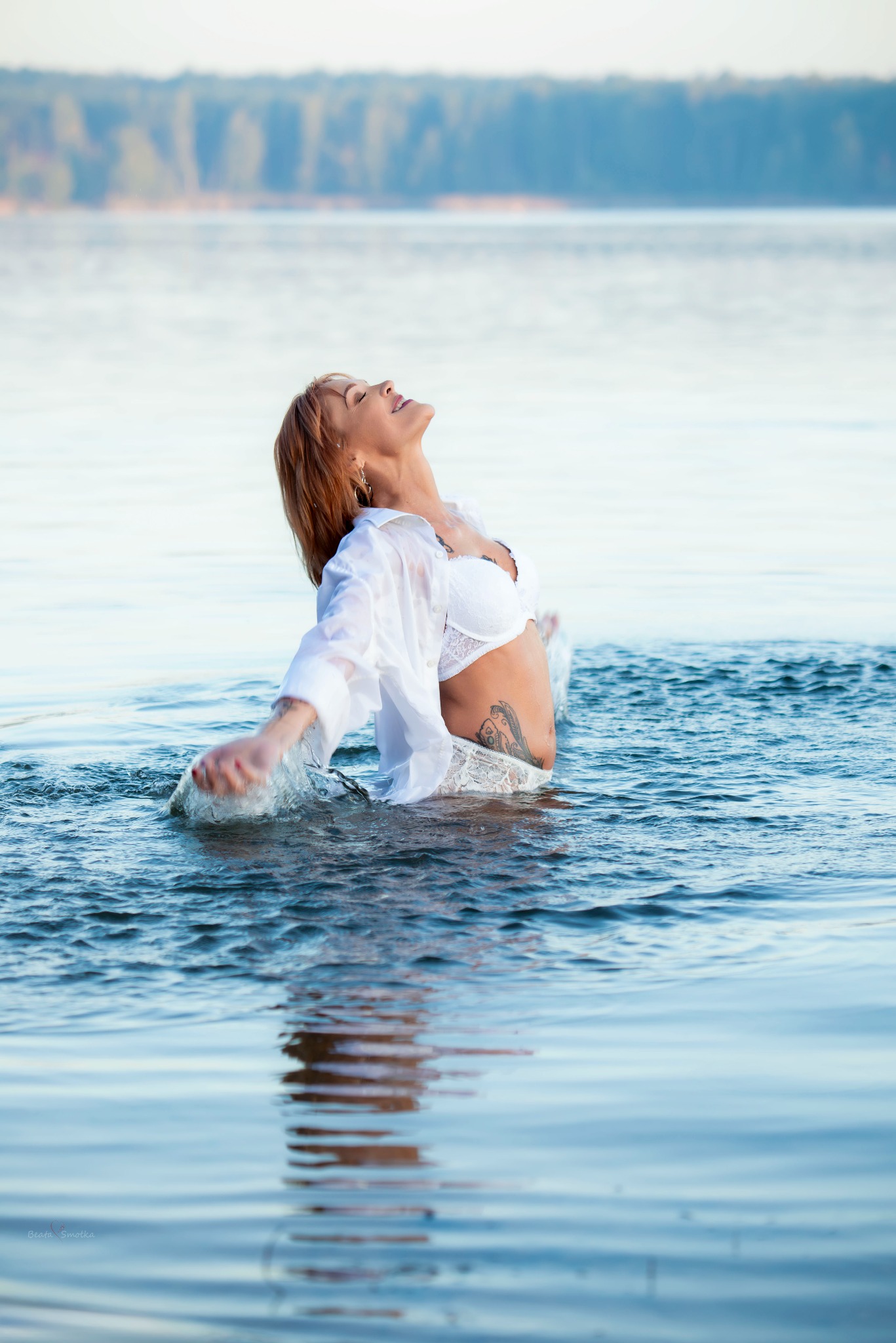 Kobieta w białej bieliźnie i koszuli zanurzona w wodzie jeziora, z tatuażem na brzuchu, odchylona do tyłu z zamkniętymi oczami, w tle las.