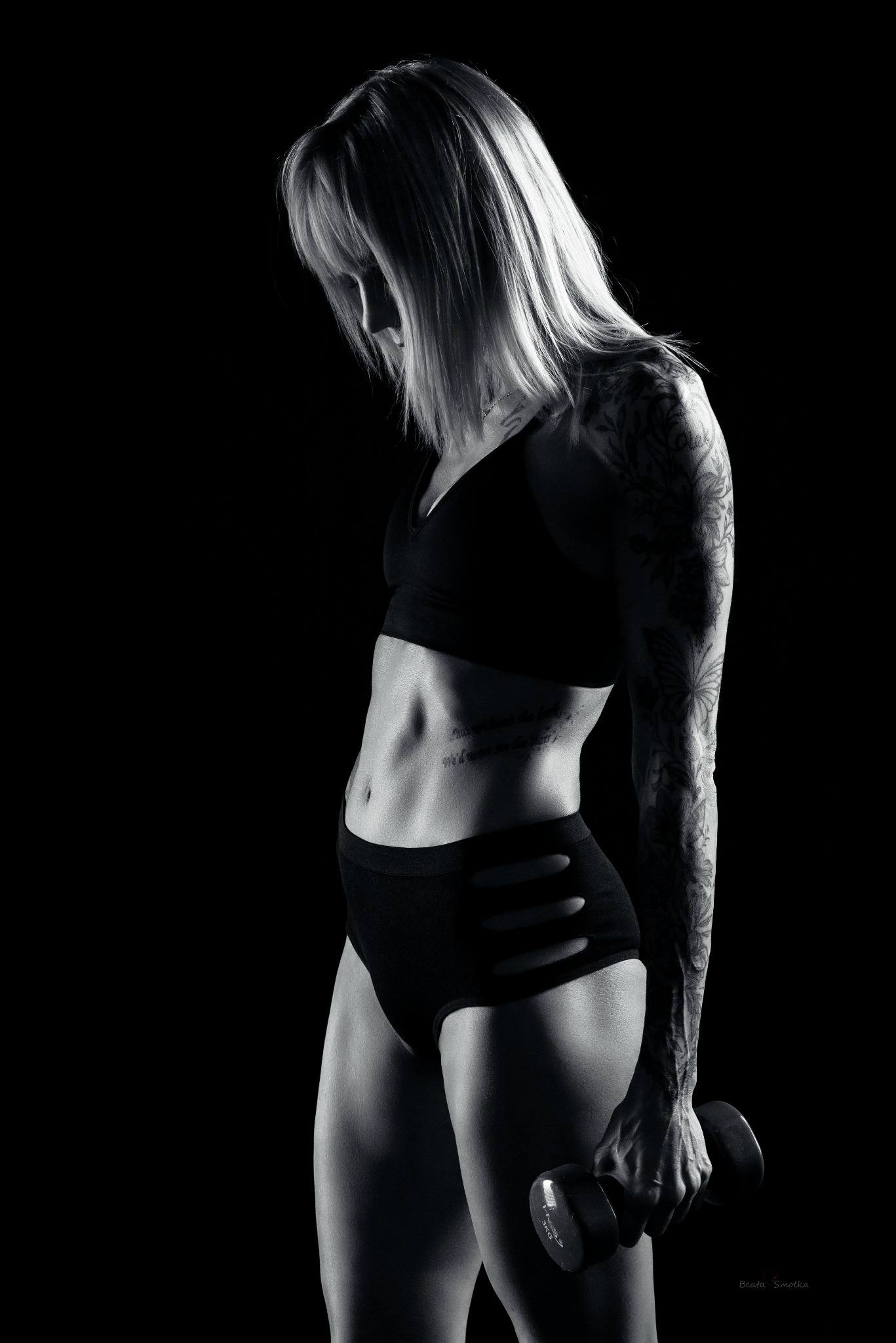 Czarno-białe zdjęcie kobiety z tatuażami, w sportowym stroju, trzymającej hantlę. Silne oświetlenie podkreśla muskulaturę i detale tatuaży na ramieniu.